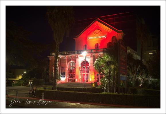 This image captures a building, the Hyères Casino, illuminated in warm red lighting at night, giving it an intense and inviting glow. The architecture reflects a historic or classical style, with large arched windows and sturdy columns that frame the entrance. The red illumination emphasizes the building’s textured facade and structural details, adding dramatic contrast against the surrounding darkness.

Above the entrance, a sign reads "Casino des Palmiers".
The surrounding area is quiet and shadowed, with silhouettes of trees and bushes softly blending with the night, lending a serene and contemplative mood. The lighting also reflects subtly on the pavement, enhancing the vividness of the scene.

Artistically, the image plays with light and shadow, creating a sense of importance and grandeur around the structure. Red as a visual theme in art often conveys intensity and draws attention, which aligns well with the building’s cultural significance.

Overall, this photograph beautifully highlights the interplay of architectural beauty and dramatic illumination, inviting viewers to appreciate the cultural space it represents.
