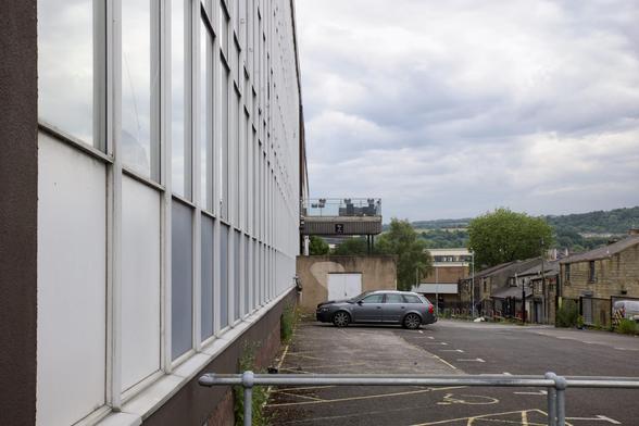 A grey car is parked in an almost empty car park next to a large building with many windows. A balcony with tables and chairs extends from the building. Small houses and green hills are visible under a cloudy sky in the background.