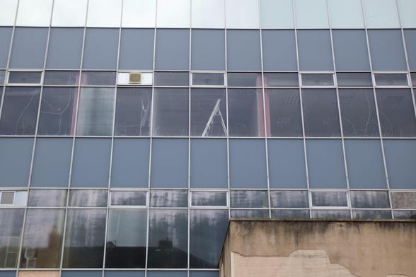A glass building facade with rectangular blue-tinted windows in a grid pattern. Some windows are foggy or streaked, and an air conditioning unit is mounted in one window. Part of an older, stained tan wall is at the bottom right.