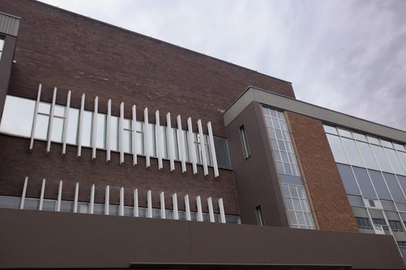 A modern brick building with large windows and vertical white metal bars on the façade. The sky is overcast, giving a muted tone to the scene. The architecture features both dark and light brown panels.