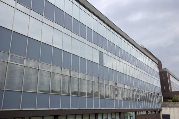 A modern, multi-storey building with large glass windows reflecting the cloudy sky. The building has a rectangular shape and blue-tinted glass panels divided by vertical and horizontal frames, creating a grid-like appearance. The structure extends into the distance.