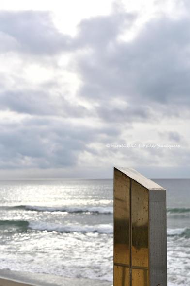 Imagen vertical donde aparece el mar y un cielo nublado. En el margen inferior derecho hay una figura de formas geométricas y apariencia cromada. Se trata del extremo superior de una ducha de playa.