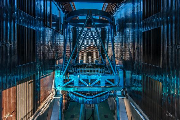 A blue toned photography of the Subaru Telescope enclosure opened in preparation for observations. Beyond the slit, Maunakea can be seen illuminated by the setting sun. 