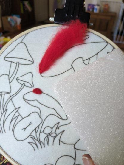 A swatch of patterned fabric with the outline of mushrooms on it, a Styrofoam block for show and red wool that's starting to be felted into the fabric.