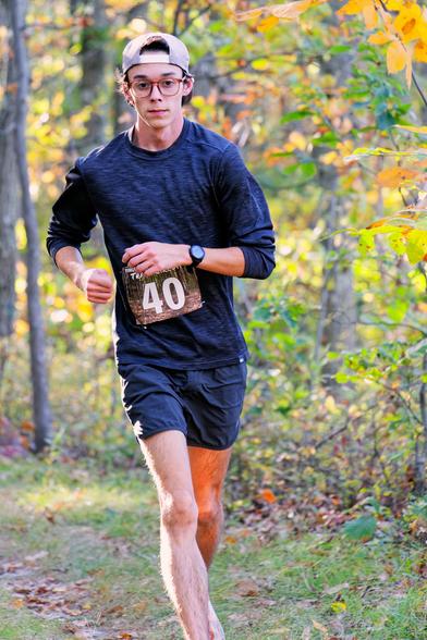 Young man in the center of the frame is running in a colorful fall color forest with a billed cap backwards, glasses, dark blue shirt and shorts, thin legs, and bib number 40