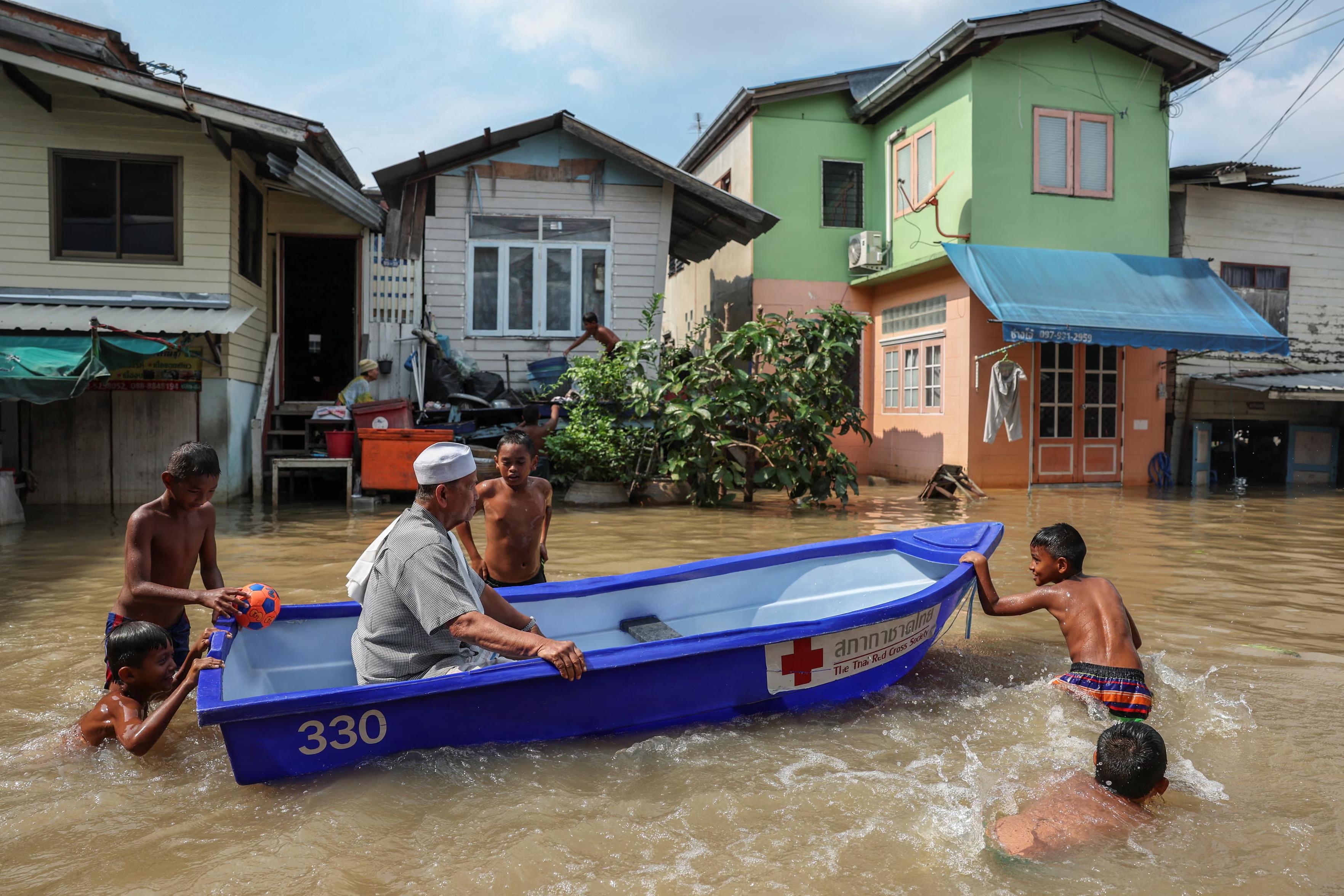 A man sits in a boat as children push it on a flooded road.