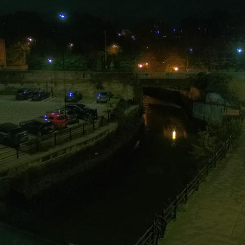 Photograph of the tidal stretch of Ouseburn River at south side of Ouseburn Bridge in Newcastle upon Tyne. To the left (south-west) of the bridge is Cut Bank, and to the right (north-east) is Byker Bank. In the foreground is the pedestrian Riverside Walk and the photo also spans some surrounding built-up areas and trees, with sky in the background. Still image captured from webcam at 03:13 hrs on Saturday 15 November 2025.