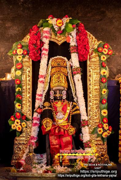Idol of Hindu deity Krishna in red standing with right palm out and turned downward (wish-granting pose), left hand on left front waist; wearing gold shoes, gold palm coverings, a 3-level gold crown, and lots of gold ornaments including a long necklace made from gold spheres. A gold arch behind Krishna. Strings of green betel leaves (alternating red and yellow flowers on them) hang down the sides of the arch. A string of white flowers (some small bands of red flowers) hangs down from the middle of the arch and flows down to the floor on both sides of Krishna. A thick string of red roses hangs at the top of the arch. Pitch black filling within the arch; dark blue background behind the arch.