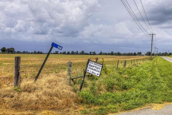 A country road stretches alongside a vast field under a cloudy sky, with road signs leaning over the grass.