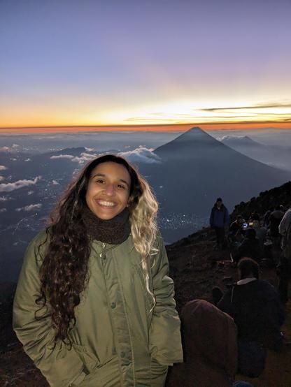 A photo of me on top of a volcano wearing a winter jacket, in the background there are a number of volcanoes and the sun is rising over them