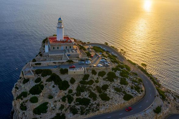Luftaufnahme eines weiß-roten Leuchtturms, der auf einer zerklüfteten, felsigen Klippe thront und majestätisch über das Mittelmeer blickt. Eine enge, gewundene Serpentinenstraße schlängelt sich den Abhang hinauf zum Leuchtturm und einem kleinen Parkplatz, auf dem Autos in der Abendsonne stehen. Rechts davon fällt die Klippe steil ins Meer ab, während auf der linken Seite die goldglänzende Oberfläche des Wassers im Licht des tief stehenden, hellen Sonnenuntergangs strahlt. Die Farben des Himmels und des Meeres sind intensiv, ein unvergesslicher Anblick.