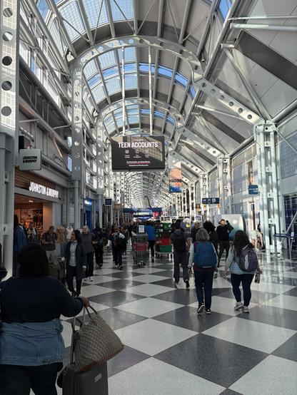 A busy airport terminal featuring modern architecture with large windows. Travelers are walking through the spacious area, which includes shops and an electronic billboard advertising “Teen Accounts.” The floor has a black and white checkered pattern, and various luggage and bags are visible.