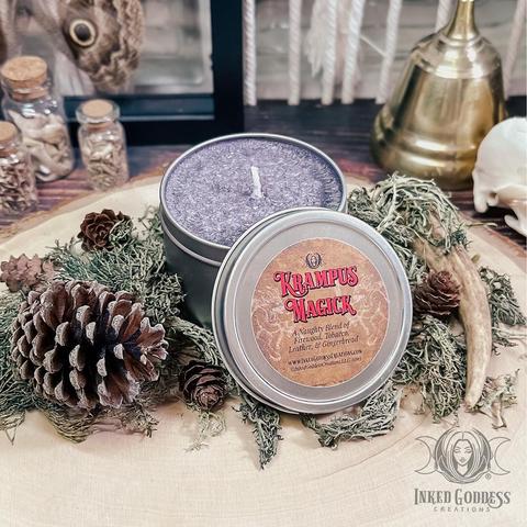 A Krampus Tin Candle is presented on a wood slice. The tin is round, wide, and silver in color, and its lid is propped up against the side. The candle is ashy purple in color. The label includes the candle's name and scent blend, firewood, tobacco, leather, and gingerbread. This is an exclusive product from Inked Goddess Creations.