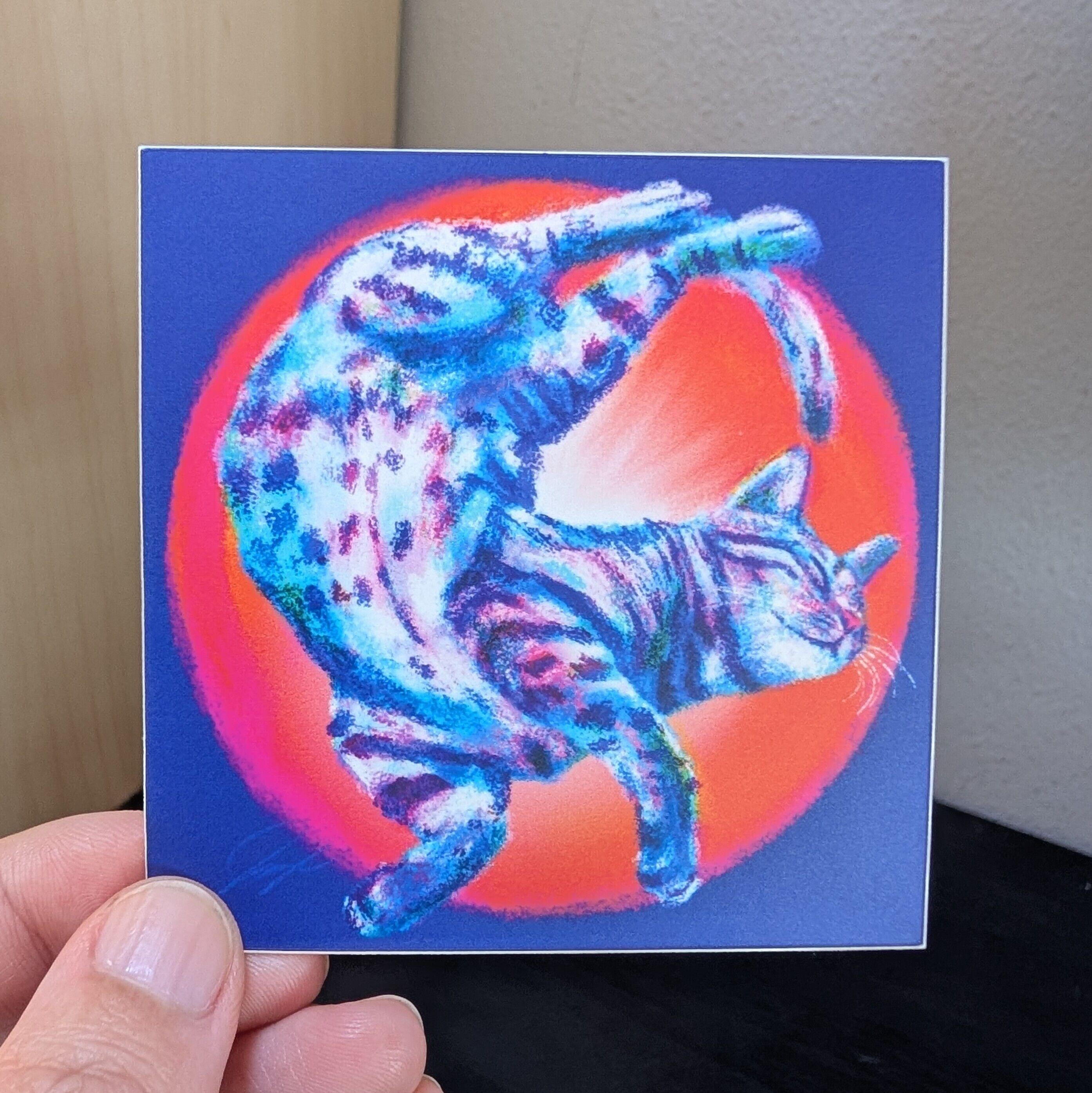 Photo of a left hand holding a square sticker. The sticker is a matte painting of a cat wiggling.