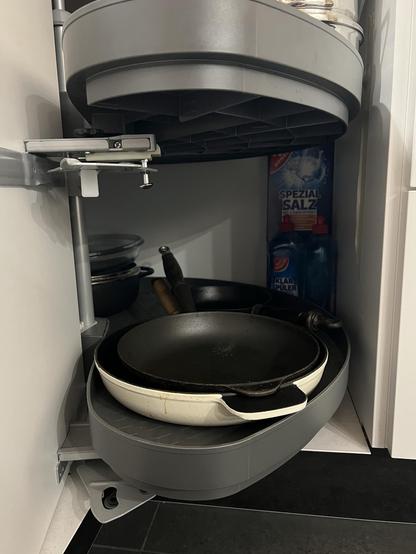 Ein offener Küchen-Eckschrank mit Rondell-Dingens für Töpfe und Pfannen. Rechts hinten in der Ecke zwischen den runden Regalböden und der hinteren Schrankecke ist etwas Platz, dort sind Kartons mit Spülmaschinen Salz und Flaschen mit Klarspüler aufbewahrt 