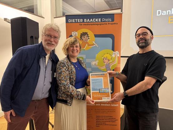 Lambert, Susan und Hannes mit der Urkunde des Preises vor einem Rollup vom Dieter-Baacke-Preis