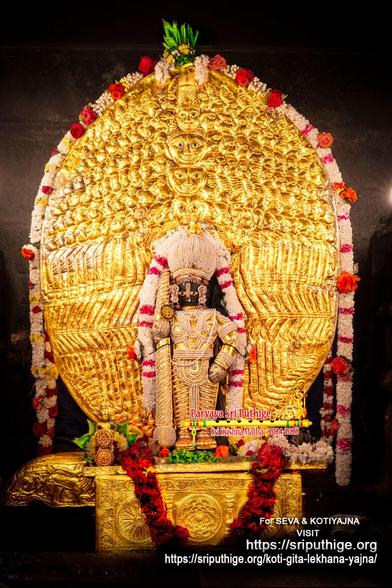 Idol of Hindu deity Krishna in pearl "chain mail" (gold undergirding), a matching dome-shaped crown, and a similarly clad staff in right hand, left hand on front left waist, standing on a gold-covered pedestal, wearing gold shoes. A string of white flowers (some bands of red flowers) around Krishna. A garland of dark red roses on the pedestal. Loose leaves of green holy basil at Krishna's feet. A large oval-shaped gilded radiant shield behind Krishna with several rows of human-like faces embossed at the top half, a row of arms in the middle portion, and a row of legs at the bottom. The embossed shapes together are designed to emanate from the main idol to illustrate a supreme being with several faces, arms, and legs.