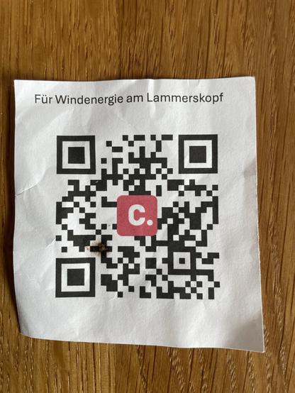 A wrinkled paper with the text "Für Windenergie am Lammerskopf" and a QR code. The background is a wooden surface.