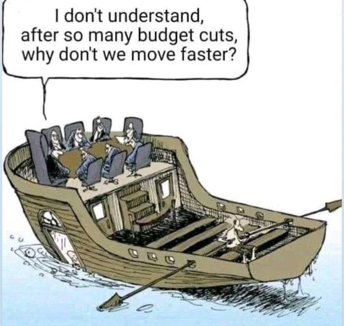 A political cartoon depicting a large boat. On the upper deck, several executives in suits sit around a meeting table, with one saying, "I don't understand, after so many budget cuts, why don't we move faster?" On the lower deck, a single, small person is struggling to row the entire boat with multiple oars.