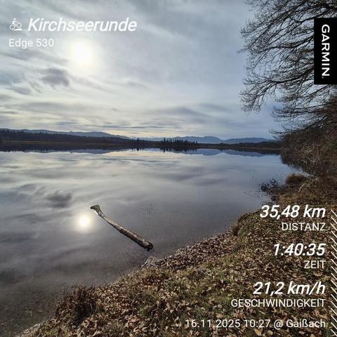 A serene lakeside scene unfolds under a soft, overcast sky, where the calm water mirrors the muted colors of the clouds and distant mountains. The lake, known as **Kirchsee**, reflects the surrounding landscape like a giant mirror, creating a sense of tranquility and stillness. A lone, weathered branch lies partially submerged in the shallow water near the shore, adding a touch of rustic charm to the scene.

The image captures a cycling route, labeled **"Kirchseerunde"** and **"Edge 530"**, hig…