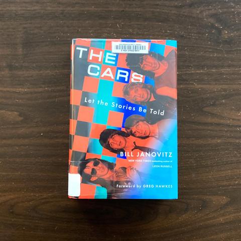 Front cover of a book titled "The Cars: Let the Stories Be Told" by Bill Janovitz. Cover shows a line-up of five white men in their 20s and 30s, of varying heights, most with shoulder-length hair, most are staring into the camera. In the background is a checkerboard pattern in blue and red.