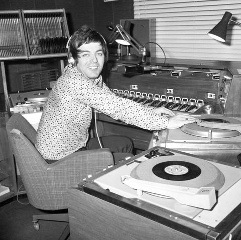 Black and white photo of a smiling male DJ (Tony Blackburn) wearing headphones in a studio operating turntables and a mixing console with numerous buttons.