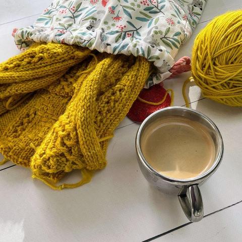 Angled view of a bright yellow knitting project spilling out of a project bag. The ball of yarn and a cup of coffee are also there on the white plank background.