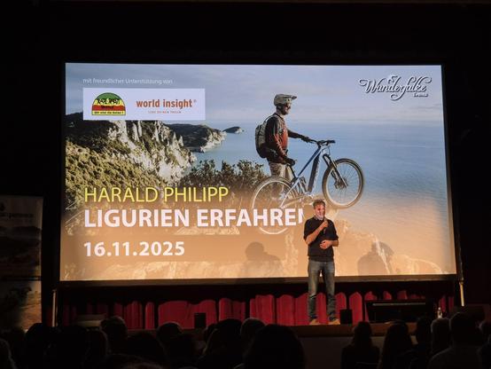 This image captures a moment from a live presentation by **Harald Philipp**, titled **"Ligurien Erfahren"** (Experiencing Liguria), held on **16.11.2025**. Harald Philipp is seen speaking on stage, with a large screen behind him displaying the event title and a stunning background image of a cyclist on a rugged coastal trail overlooking the sea.

The screen also features logos for **"Wunderfälke Events"** and **"world insight"**, indicating the event's friendly support and focus on inspiring tr…