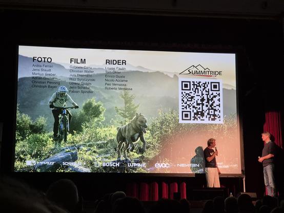 This image captures a lively stage presentation, likely at a mountain biking or outdoor adventure event. The large screen behind the speakers showcases a dynamic scene featuring a mountain biker and a dog running through a scenic trail, symbolizing adventure and companionship in the great outdoors.

The screen is divided into three sections, each listing notable individuals in the fields of **Foto (Photography)**, **Film**, and **Rider**:
- **Foto**: Ardita Ferrari, Jens Staudt, Markus Greber, …