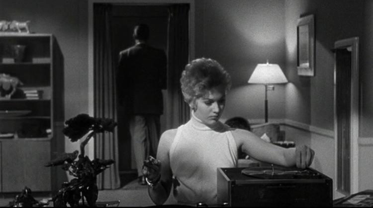 Black and white scene featuring a blonde woman in a white turtleneck operating a record player while a man stands in a doorway in the background.