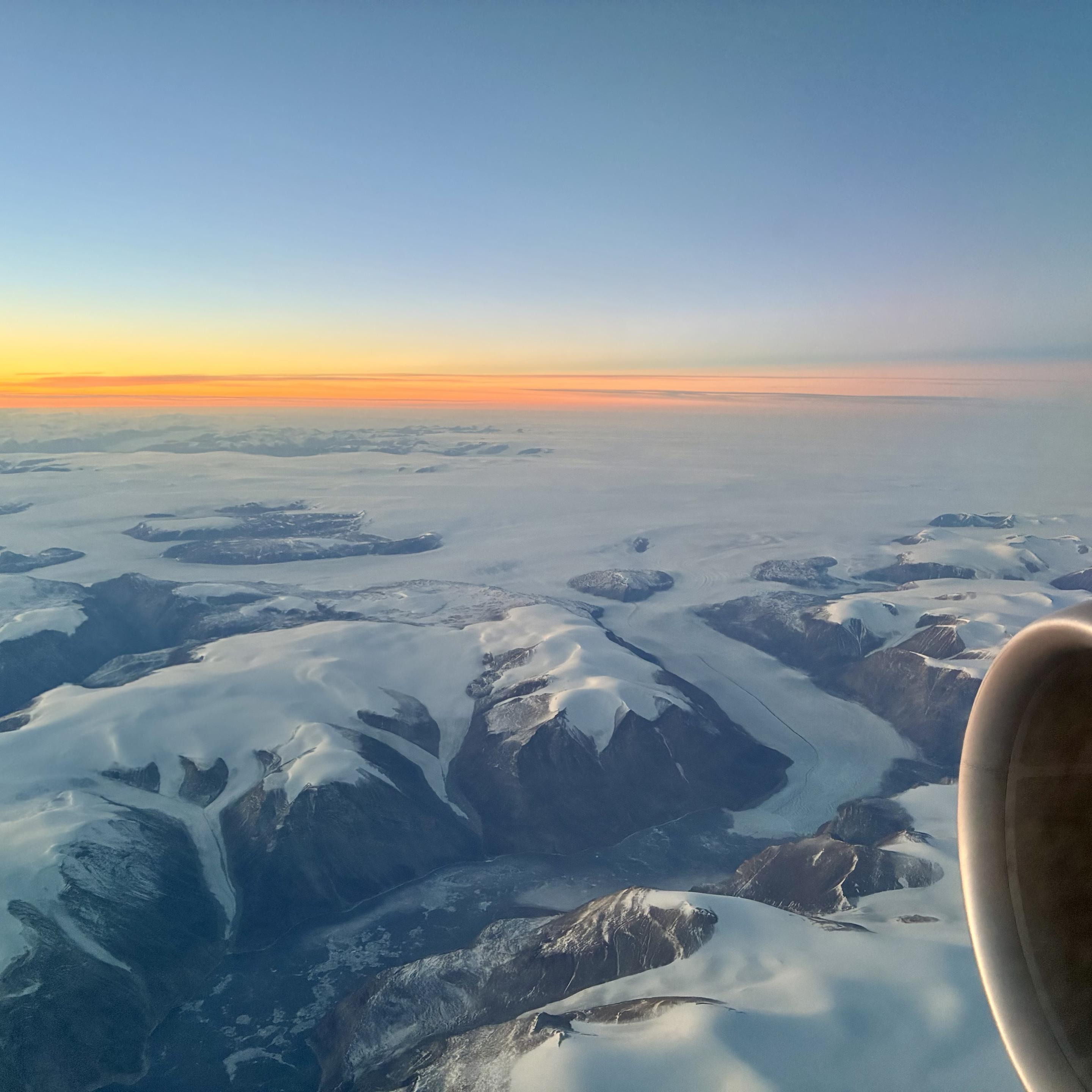 Aerial view of snow-capped mountains and glaciers under a clear sky at sunrise, with a soft orange horizon and an aircraft engine visible in the corner.