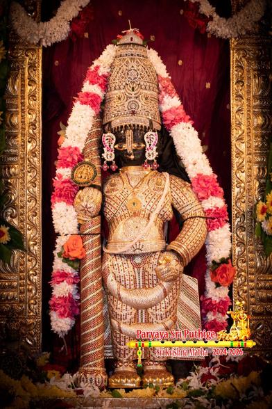 Idol of Hindu deity Krishna in diamond cladding with a matching crown holding a similarly clad staff in right hand. A string of white flowers (some bands of red roses) around Krishna. The sides of a gold arch, under which Krishna is situated, are partly visible. Red cloth background behind the arch.