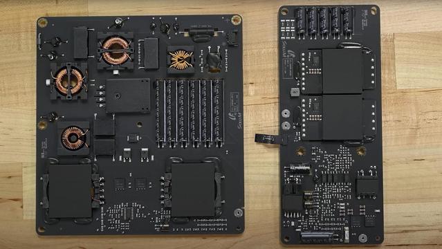 High-resolution photograph of two internal printed circuit boards from the power supply module of the Apple Studio Display. On the left is a larger black board featuring multiple toroidal and bobbin wound inductors, a bank of electrolytic capacitors aligned vertically, and two large transformer blocks clad in black heat-shielding. On the right is a narrower board, also black, showing several high-current MOSFET power stages covered with black shielding blocks, smaller capacitors at the top, and numerous surface-mount components in the lower section. Both boards are mounted on a light wood surface, revealing mount holes at the corners and labelled connectors on the right board.