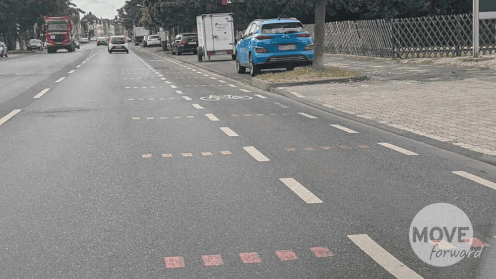Foto eines Fahrbahnabschnitts mit Radschutzstreifen. auf und vor allem neben dem Schutzstreifen sind Punktlinien auf den Asphalt gemalt, mit denen Seitenabstände "nach Augenmaß" leicht gemacht werden