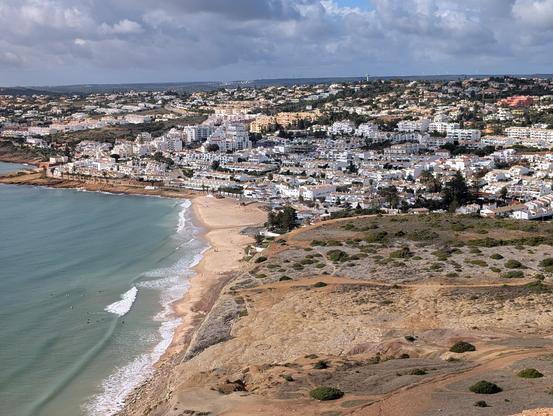 Top half is a city of white buildings along a hill against blue sky. Bottom left is a turquoise sea washing up on a beach. Bottom right is a brown plateau with scrubby green plants. 