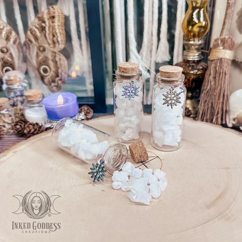 Three specimens of the "Snow Quartz in Jar" product are arranged on a raw wood slice. The small vials are clear and have cork stoppers with an iridescent snowflake charm tied around the neck. They contain several small rough pieces of opaque white quartz. One vial is tipped over and the quartz pieces are arranged just outside the uncorked opening. Presented by Inked Goddess Creations.