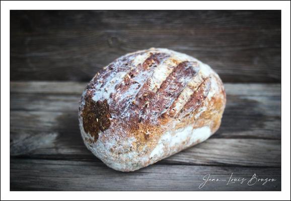 
The image captures a rustic loaf of bread resting on a wooden surface, inviting a sensory appreciation of its texture and form. The bread's crust boasts a beautifully browned, artisanal finish with a pattern of diagonal slashes across the top, likely made to help the loaf expand during baking. These slashes add a rhythmic visual interest and hint at a careful, handcrafted process.

The crust appears robust yet slightly floured, creating a contrast between the darker baked sections and the lighter dusted areas. The subtle flour dusting evokes the traditional, old-world approach to bread-making, suggesting this loaf is likely sourdough or another type of naturally leavened bread.

The wooden backdrop, with its weathered grain and muted tones, complements the loaf’s organic character, enhancing a sense of warmth and homeliness. This setting evokes cultural significance related to the age-old craft of baking—symbolizing sustenance, simplicity, and nurturing care.

Overall, the composition is a quiet celebration of wholesome, artisanal food, grounded in tradition and crafted with patience. It visually conveys comfort, nourishment, and the timeless art of bread-making.
