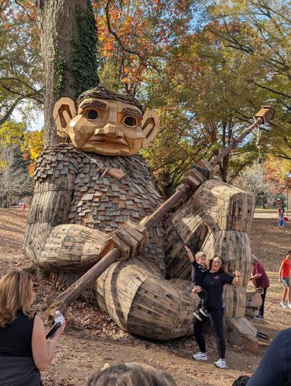 A 18 feet wood sculpture of a troll sitting on the floor with his back on a tree