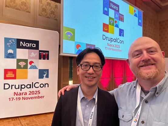 Two attendees smiling in a conference hall at DrupalCon Nara with a large screen and banner that read “DrupalCon Nara 2025, 17–19 November.”
