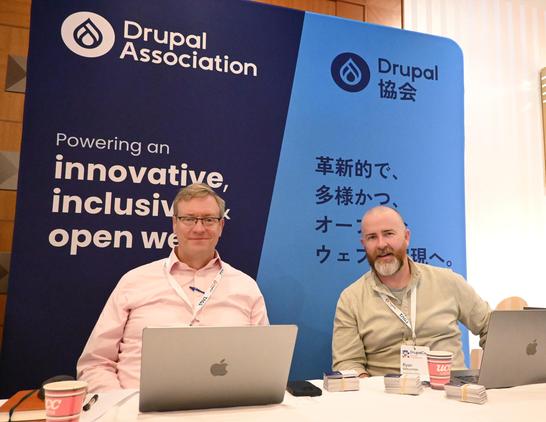Tim Doyle and Ryan Witcombe sitting at a booth with Drupal Association backdrop behind them.