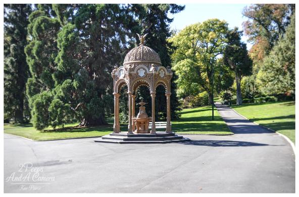 A photograph of a historic, highly ornate rotunda structure situated in a well maintained park.

The rotunda has a domed roof topped with a sculpture and features several arches supported by slender pillars, with a small central fountain visible inside.

It is surrounded by lush green lawns and large, tall trees, some dark green and some bright green, indicating a sunny day. A paved pathway curves away to the right of the rotunda, leading up a gentle slope.