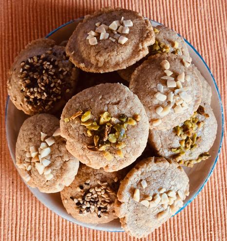 Runde Kekse, bestreut mit Pistazien-Schredder, Mandel-Splittern und Sesam. Die Kekse sind hellbraun und machehn einen nahrhaften Eindruck. Sie liegen auf einem runden Teller (der eine Unertasse ist, nur zum Größenvergleich) und dieser kleine Teller steht auf einem orange-farbenen Tischset, das gerippt ist.