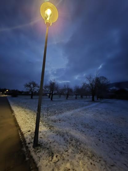 A serene winter evening unfolds under a deep blue sky, where a solitary streetlamp casts a warm, golden glow over a snow-dusted park. The light from the lamp illuminates the immediate area, creating a striking contrast between the bright foreground and the shadowy, snow-covered field that stretches out into the distance. The snow, lightly blanketing the ground, adds a sense of calm and stillness to the scene.

In the background, bare trees stand tall, their silhouettes stark against the twiligh…