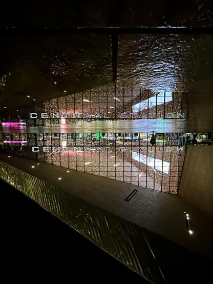 A modern train station, labeled "CENTRAAL STATION," is depicted with a reflective surface showing colorful lights and glass architecture.