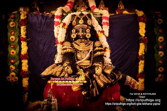 Idol of Hindu deity Krishna in black (broad gold embroidered border) appears as an infant crawling/running; seat covered in red cloth; wearing gold shoes, gold palm covering, and lots of gold ornaments. A string of white flowers (some red and yellow flowers) around Krishna waist-waist. Dark blue (almost indigo) background. A string of white flowers (some red flower bands) hangs from the ledge in the background behind Krishna. Strings of yellow flowers hang at the edges of the background. Strings of green betel leaves (with yellow and red flowers on top) also hang at the edges.