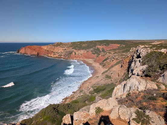 A totally blue sky, rocky brown-red cliffs with short scrubby vegetation on top, leading to a beach with choppy turquoise waves. 