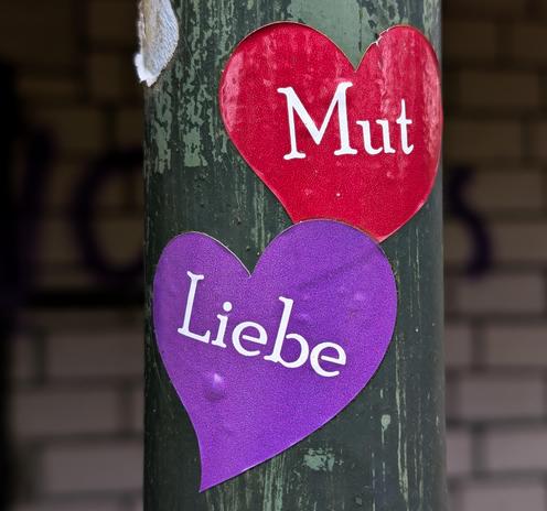 Zwei Aufkleber auf einem Pfahl.  "Liebe" in lila und darüber "Mut" in rot.