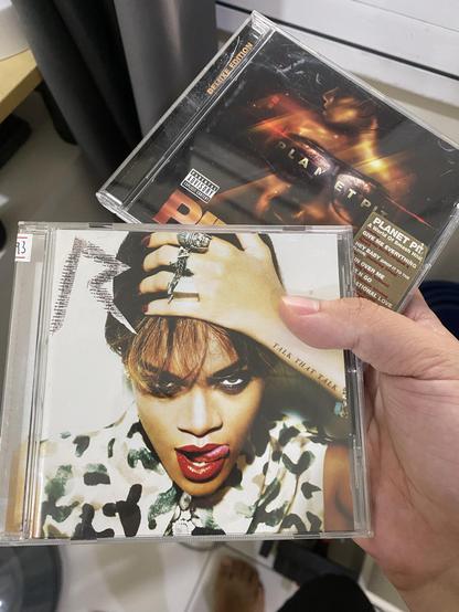 A hand holding two music CDs. The front CD features a close-up cover of a woman with red lipstick, camo-patterned clothing, and her tongue slightly out (Rihanna’s Talk That Talk album). Behind it is another CD with a darker, fiery-toned cover showing a stylized face and the text “Planet Pit (Deluxe Edition) (Pitbull’s Planet Pit-Deluxe Edition album).”