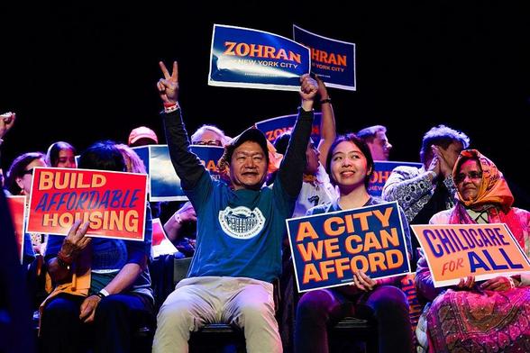 A crowd with some prominent east asian and south asian people are holding signs like BUILD AFFORDABLE HOUSING, A CITY WE CAN AFFORD, CHILDCARE FOR ALL, ZOHRAN: FOR NEW YORK CITY