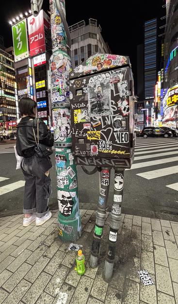 nighttime shot across the road from kabukicho; a girl waiting to cross the road, and next to her an electrical/signal box (covered in stickers and graffiti) standing on two slightly bent thin metal poles that look like legs, making the box also look like it's waiting to cross